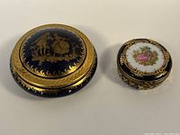 Two Limoges porcelain trinket boxes side by side. Larger cobalt blue round box with gold courting scene and smaller oval box with painted courting couple under glass lid with gold trim.