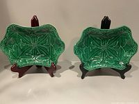 Two Wedgwood Majolica green leaf plates on display stands, side by side. The plate on the left has visible chips; the plate on the right is in better condition.