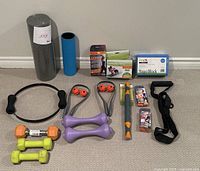 Photo showing all items in the Pilates and weights lot arranged on carpet against wall. Items include foam rollers, Pilates ring, hand weights, yoga block, massage rollers, resistance bands, and exercise grips.