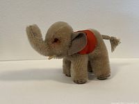 Side view of tan mohair miniature elephant toy with trunk up, visible red felt saddle, grey felt ears, and cord tail.