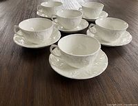 Set of six white bone china cups each paired with a matching embossed saucer featuring the cabbage leaf design on a wooden table.