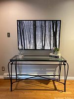 Full view of console table with glass top and metal legs under black and white forest scenery wall art.
