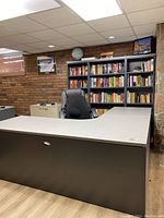 Full view of grey L-shaped desk in office setting with bookshelves behind it and black office chair positioned in front.