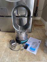 Front view of Dyson AM10 humidifier with transparent water tank, magnetic remote, power cord and manual on floor