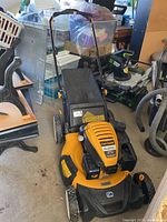 Front view of Cub Cadet mower showing yellow-orange deck, black handle and attached grass bag in garage setting