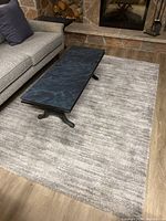 Grey textured carpet with rectangular black coffee table on top, placed in front of a grey sofa and stone fireplace.
