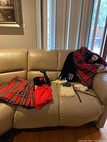 Overall view of Highland dancer costume pieces on beige couch including tartan kilt, red shorts, black bonnet hat, lace cuffs, and white tassels.