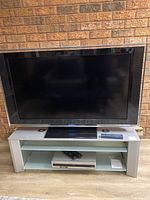 Front view of Sony TV mounted on grey console with green glass shelves, showing DVD/VCR player and remotes