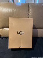 Closed UGG shoe box placed on sofa, showing UGG logo on top.