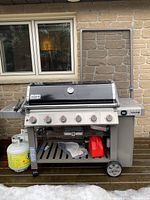 Front view of assembled Weber Genesis II grill on deck with propane cylinder attached and accessories under shelf.