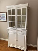 Front view of white wooden cabinet showing glass-paned upper doors, two drawers, and lower cabinet doors.
