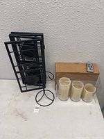 Three ivory battery powered candles in plastic packaging alongside a cardboard box and three black metal candle holders including a wall mounted triple holder and freestanding holders, plus a remote control on the box.