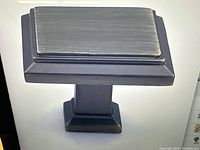 Close-up photo of a single square black matte cabinet knob showing the flat rectangular top and square stem and base.