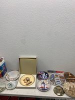 Full view of lot showing various collectible plates with floral, folk art, lighthouse, and scenic motifs, along with a souvenir tray and coasters.