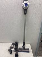 Dyson V6 vacuum assembled with long metal stick and motorized floor head, wall charging dock, charger, and brush attachments on a white table.