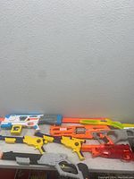 Photo of seven toy blasters arranged on a white surface against a plain wall, showcasing a variety of colors and designs.