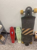 Three skateboards leaning against a wall including one large longboard with black deck and two smaller plastic cruiser boards, red and light green.