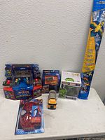 Full lot showing various toys including Akedo Battle Arena, Hot Wheels set, Marvel Hulk Funko Pop, Spider-Man mask, yellow toy truck, and parrot kite on white table with gray wall background.