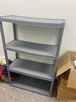 Full view of gray plastic storage rack with four shelves, showing the overall structure and condition against a wall.