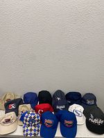 View of 16 sports hats arranged in rows on a floor against a wall, showing various colors and logos.
