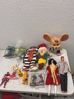 Photo showing assortment of vintage toys including plush-like dolls, large cartoon mouse figurine, mini animal figurines, and action figures on a white surface.