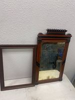 The two items side by side: a carved wood framed mirror with partial wear on the mirror glass and a plain wooden rectangular frame.
