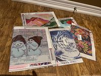 Five diamond painting canvases laid on floor showing designs of snowmen, floral art, Santa Claus, and landscape.