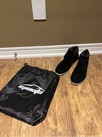 Black shoes placed on a wooden floor next to a black nylon drawstring bag with white print.
