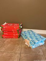 Photo showing 5 stacks of red and green packaging of Staples copy paper 8.5x14 legal size and 2 stacks of blue packaging for multipurpose tan paper 11x17 size.