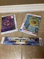 Three diamond art craft kits on tiled floor, two in sealed packaging and one rolled canvas with colorful designs.