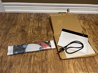 Diamond art canvas in sealed packaging alongside diamond art light board and USB cable in an open box on wooden floor.