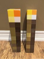 Two pixelated Minecraft torch replicas standing upright against a wall showing their front faces: blocky colors in yellow, orange, white on top and shades of brown below.