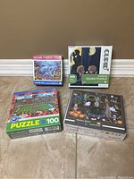 Four boxed jigsaw puzzles arranged on floor against brown wall: Dolphins Love, couples silhouette, soccer stadium 100 pieces, Mystery Witch theme.