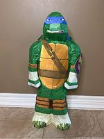 Front view of Teenage Mutant Ninja Turtle piñata, showing green turtle body, blue mask, yellow chest, brown straps, and white feet.