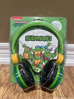 Front view of Teenage Mutant Ninja Turtles kids headphones in original sealed packaging, showing green headband and turtle character artwork.