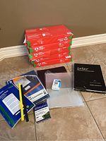 Overview of five packs of paper with additional miscellaneous office supplies on the floor near the paper stack including document folder, sheet protectors, binder clips container, foil sheets pack, envelopes