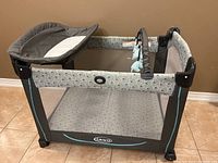 Full view of Graco portable playpen showing gray and teal color, mesh sides, attached padded changing table, and toy bar with hanging toys