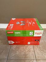 Closed Staples box containing 10 packs of legal size printer paper on tile floor.