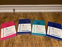 Four sealed Go Girl Budget Planner books laid flat, colors pink, dark blue, light blue, and purple visible, showing front covers with title and budget planner details.