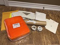 View of assorted envelopes, orange bubble mailers sealed in package, brown kraft packaging wrap roll, packing tapes and envelopes arranged on floor.