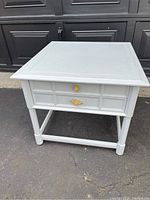 Front-side angle view of a white painted side table showing two drawers each with gold-colored handles, four legs with stretcher.