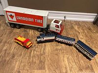 Metal Tonka Canadian Tire semi-truck with white trailer featuring Canadian Tire logo, accompanied by a small fire truck and wooden train cars on wooden floor.