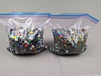 Two large clear resealable plastic bags full of assorted glass marbles with different colors and designs placed side-by-side on a grey fabric surface.