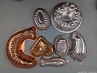 Photo showing all seven different metal cake moulds in a flat lay on a gray surface, featuring copper and tin materials with vintage embossed decorative patterns.