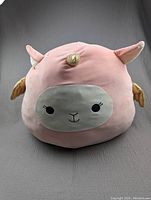 Front-facing view of the pink unicorn Squishmallow showing the white embroidered face with eyes, nose, gold horn, pink ears, and gold wings.