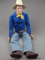 Full body of the John Wayne doll seated, showing blue shirt, light blue pants and brown boots along with white cowboy hat and yellow neckerchief.