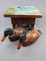 Color photo showing the painted wooden stool with a nature lake scene painted on the seat, and two wooden ducks lying in front of it.