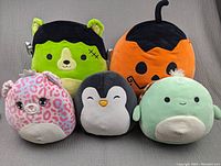 Five Squishmallows plush toys shown on a gray fabric background. Characters include a Frankenstein bear, orange pumpkin, pink leopard cat, black and white penguin, light green round character with small arms.