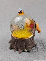 Side view of the Winnie the Pooh musical snow globe with Pooh inside the glass globe reaching toward the honeycomb base. The base resembles a tree stump with small flowers. A bee rests on top of the globe.