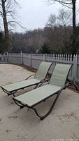 Two outdoor chaise loungers positioned side by side on a patio with fenced background, showing entire loungers and sleeping surfaces.